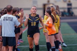 Girl in soccer uniform waves during a team lineup, coach encourages players in the background on the field.