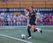 Two female soccer players compete for the ball on a field, with teammates watching from the sidelines.