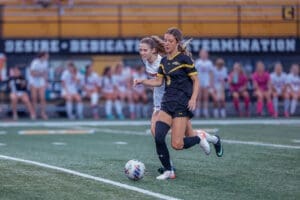 Two female soccer players compete for the ball on a field, with teammates watching from the sidelines.