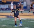 A female soccer player in a black jersey kicks a soccer ball on a field during a game. Teammates are in the background.