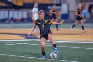 A female soccer player in a black jersey kicks a soccer ball on a field during a game. Teammates are in the background.