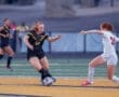 Two female soccer players compete on the field; one dribbles the ball while the other tries to intercept.