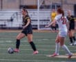 Two female soccer players compete on the field, one dribbling a ball while the other closely defends.