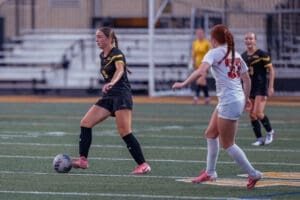 Two female soccer players compete on the field, one dribbling a ball while the other closely defends.
