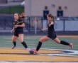 Two women playing soccer on an outdoor field, one dribbling the ball while the other supports her teammate.