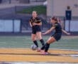 Two female soccer players in black uniforms are playing on a field, one kicking the ball while the other looks on.