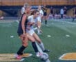Two female soccer players competing for the ball on a field, with teammates in the background.