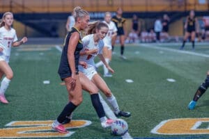 Two female soccer players competing for the ball on a field, with teammates in the background.