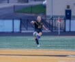 Female soccer player in a black jersey kicks a soccer ball on a turf field during a match.