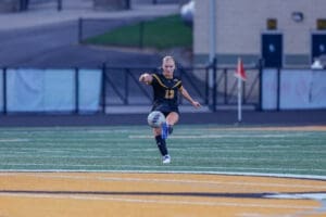 Female soccer player in a black jersey kicks a soccer ball on a turf field during a match.