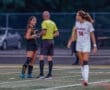 Girls' soccer match with players interacting with the referee on the field during an evening game.