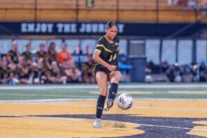 Female soccer player in black uniform kicking a ball on the field, with teammates in the background.