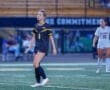 A female soccer player in a black jersey walks on the field during a game, focused on the match ahead.