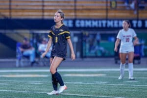 A female soccer player in a black jersey walks on the field during a game, focused on the match ahead.