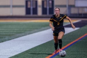 Young female soccer player in a black jersey running on the field with a soccer ball during a game at night.