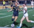Two female soccer players competing for the ball during a night game on a well-lit field. Intense athletic action captured.