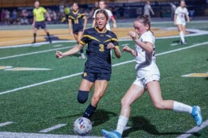 Two female soccer players competing for the ball during a night game on a well-lit field. Intense athletic action captured.
