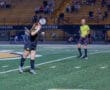 A female soccer player heads the ball during a night game, with a referee observing in the background.