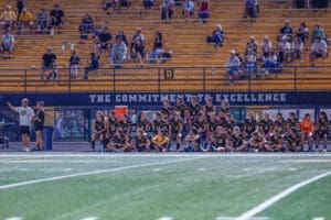 Team huddled on the sidelines during a sports event, with spectators in the background, showcasing school spirit and unity.