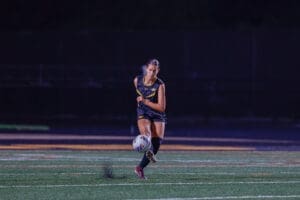 Young female soccer player kicking a ball on a field during a night game, showcasing athleticism and skill.