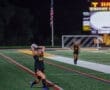 Player in black jersey throws the ball in during a night soccer game at Wright Field, with scoreboard and flag visible.