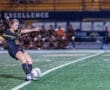 A female soccer player in action, striking the ball on a night field, with teammates visible in the background.