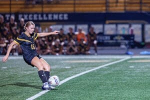 A female soccer player in action, striking the ball on a night field, with teammates visible in the background.