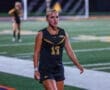 A focused female soccer player in a black uniform walks confidently on a field during an evening match.