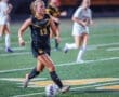 A female soccer player in a black uniform dribbles a ball on the field, focused and in motion during a game.