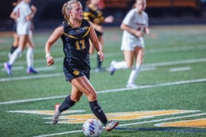 A female soccer player in a black uniform dribbles a ball on the field, focused and in motion during a game.