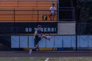 A soccer player performs a kick on the field, with spectators watching in the background. Go Tigers! visible.