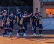 Soccer players celebrating a goal, wearing black and gold jerseys, on a field during a team match.