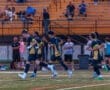 Youth soccer players in team uniforms practice running on the field, with spectators watching in the background.