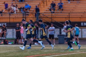 Youth soccer players in team uniforms practice running on the field, with spectators watching in the background.