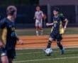 High school soccer player in black and yellow uniform dribbles the ball on a field during a night game.