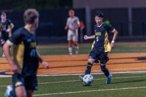 High school soccer player in black and yellow uniform dribbles the ball on a field during a night game.