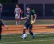 A player in a black soccer uniform dribbles the ball on a field during a night game, with teammates in the background.