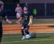 Soccer player in black jersey dribbles the ball on a field during a night game, focused on the action ahead.