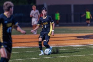 Soccer player in black jersey dribbles the ball on a field during a night game, focused on the action ahead.