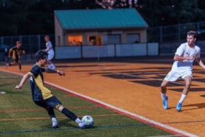 A soccer player in a yellow and black uniform kicks a ball as an opposing player runs nearby during a competitive match.