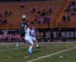 Two soccer players compete for the ball during a game, with spectators watching in the background.