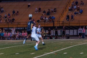 Two soccer players compete for the ball during a game, with spectators watching in the background.
