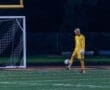 Goalkeeper in a yellow uniform prepares to play with a soccer ball on a field at night under stadium lights.