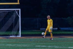 Goalkeeper in a yellow uniform prepares to play with a soccer ball on a field at night under stadium lights.