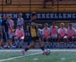 Soccer player dribbling the ball during a game, with teammates seated on the sidelines in the background.