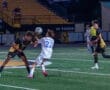 Two soccer players contest the ball on a well-lit field, with spectators in the background. Night game action shot.