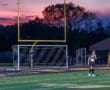 High school soccer players on a field at sunset, with goalposts and colorful sky in the background.