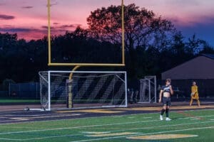 High school soccer players on a field at sunset, with goalposts and colorful sky in the background.