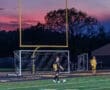 Two young soccer players on a field at dusk, with a colorful sunset and goalposts in the background.
