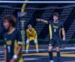 Player wearing a Tigers soccer jersey points on the field during a night game, with teammates and goalpost in the background.